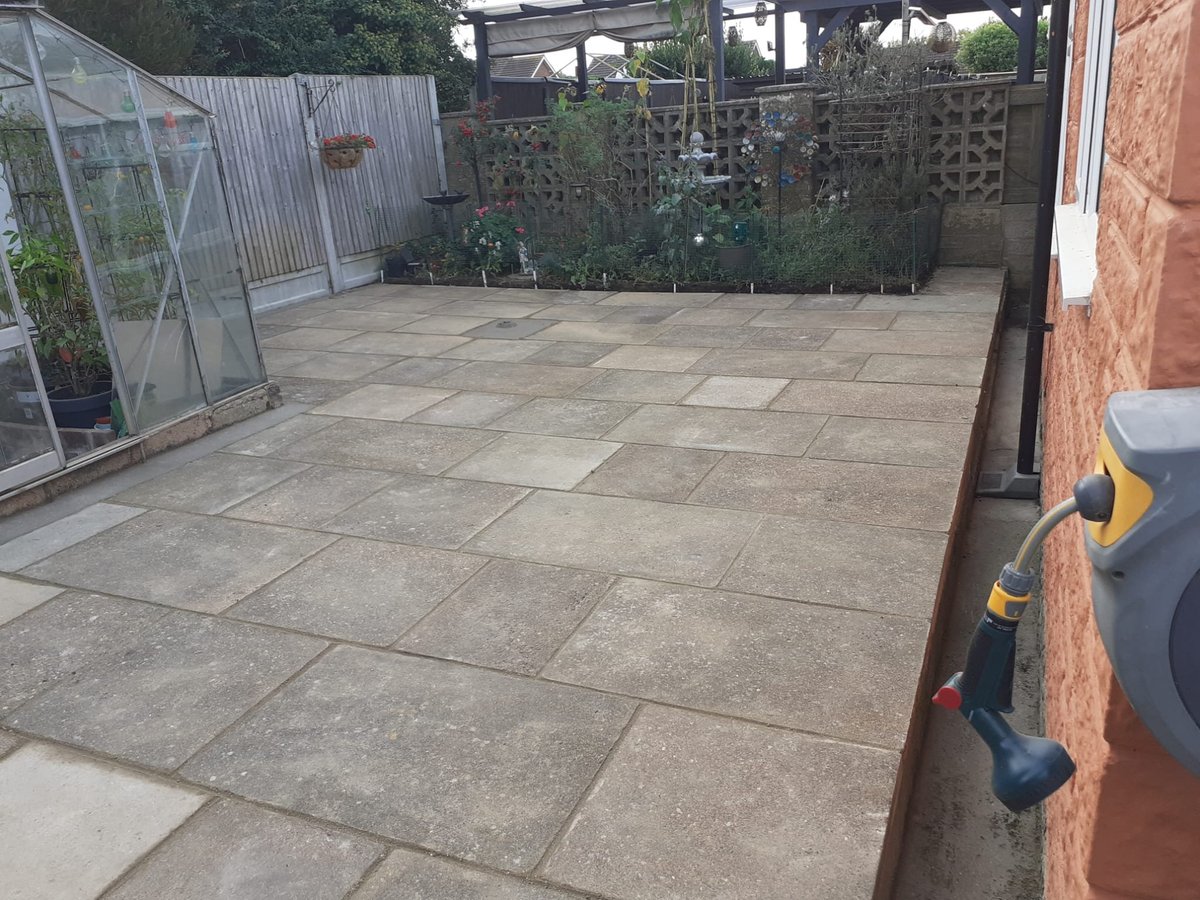 Empty patio with gray stone paving, greenhouse structure on left, brick wall on right, and overgrown fence in background